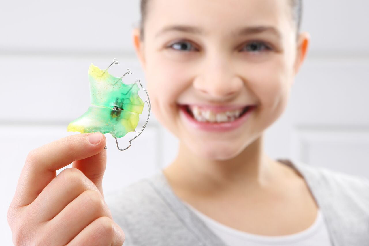 Young girl holding her yellow and green Hawley retainer, stressing the importance of long-term orthodontic success.